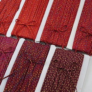 以下が含まれることがあります： メタリックな光沢のある赤とボルドーの刺繍糸が6個の糸巻きに巻かれています。糸は白い厚紙の糸巻きに巻かれており、各糸巻きの真ん中にはリボンが結ばれています。