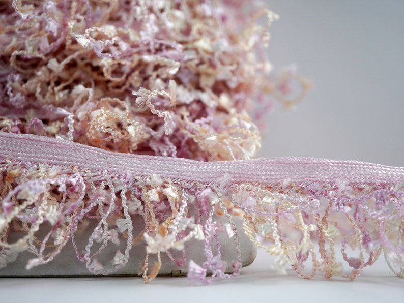 Puede incluir: Borde de flecos rosa y crema con una textura delicada. El borde tiene una base tejida y un fleco esponjoso.