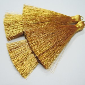 May include: Two gold tassel trims with a metallic sheen. The tassels are made of thin, silky threads and have a loop at the top for attaching.