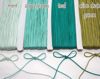 May include: Close-up of four spools of thread in shades of green: mint, aqua green, teal, and olive drab green. Each spool is labeled with its color name. Below each spool is a tied bow made from the corresponding thread.