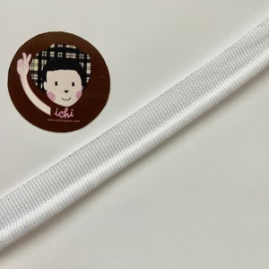 May include: White bias tape with a central crease, angled across a white surface. A circular sticker with a cartoon illustration of a person with a peace sign hand gesture and the text "ichi" is in the upper left corner.