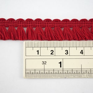 May include: Red fringe trim measuring 1 inch wide, shown against a ruler. The fringe is made from a soft, woven fabric.