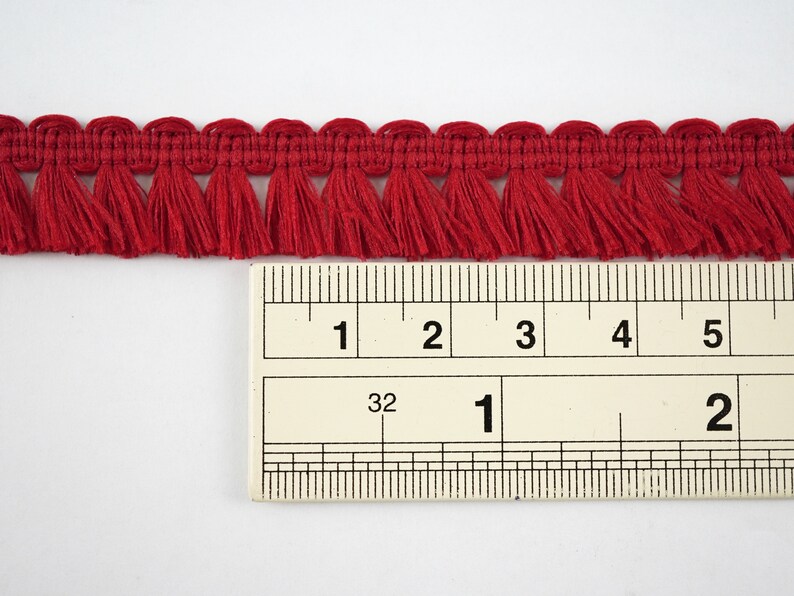 May include: Red fringe trim measuring 1 inch wide, shown against a ruler. The fringe is made from a soft, woven fabric.