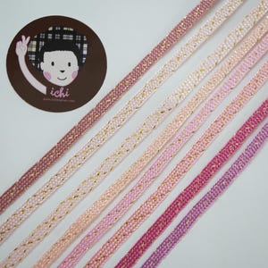 May include: An assortment of pink and gold beaded trim ribbons in various shades, arranged diagonally. A circular sticker with a cartoon illustration and the word "ichi" is in the upper left corner.