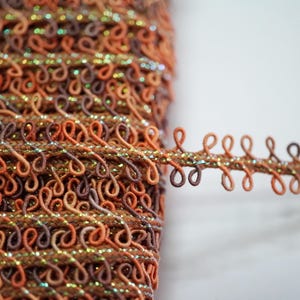 Op de afbeelding: Een spoel met bruine en oranje bies met een glinsterende afwerking. De bies is gemaakt van kleine lussen en is perfect om elke project een vleugje glans te geven.