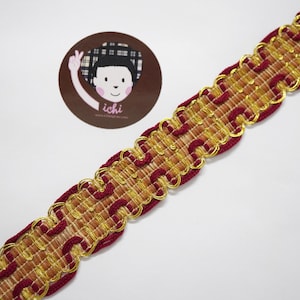 May include: Woven decorative trim in gold, burgundy, and brown. The trim has a scalloped edge with gold metallic detailing. A circular sticker with a cartoon illustration and the word "ichi" is visible in the background.