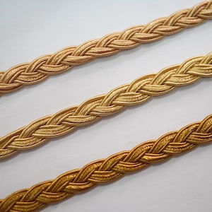 May include: Three braided cords in varying shades of gold. The cords are tightly woven together.