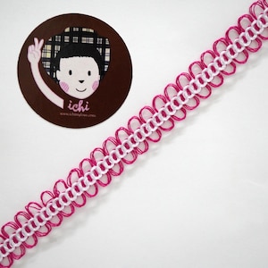 Puede incluir: Un adorno decorativo con un patrón repetitivo de bucles rosas y un centro blanco. El adorno está en ángulo sobre una superficie blanca, con una pegatina circular en la esquina superior izquierda con un personaje de dibujos animados y el texto "ichi".