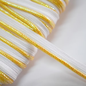May include: Close-up of a spool of white and gold ribbon. The ribbon has a flat, woven texture with a white center and a shimmering gold edge. The ribbon is tightly wound, with a portion unspooled.