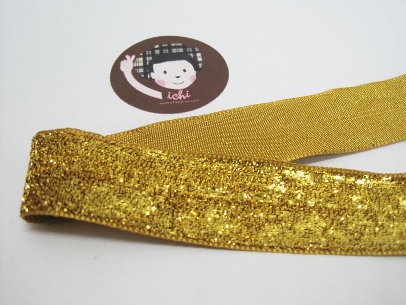 May include: Gold glitter ribbon with a subtle shimmer. The ribbon is folded in half and has a textured surface.