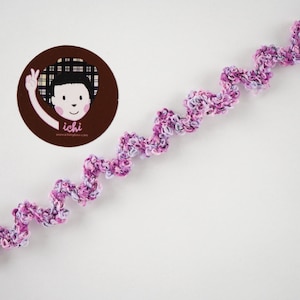 Puede incluir: Un adorno decorativo en forma de espiral en tonos de rosa, morado y blanco. El adorno está hecho de un material suave y texturizado y está colocado sobre un fondo blanco. Un logotipo circular con una ilustración de dibujos animados y el texto "ichi" está en la esquina superior izquierda.