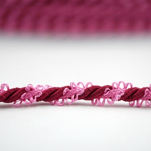Pode incluir: Close-up de um cordão torcido com guarnições rosa e bordô. O cordão é feito de um material macio e flexível e tem uma borda decorativa com franjas.