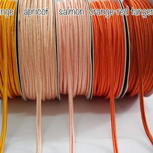 Puede incluir: Cinco carretes de cordón en diferentes tonos de naranja, desde un amarillo brillante hasta un rojo anaranjado oscuro. Los carretes están etiquetados como "naranja", "albaricoque", "salmón", "naranja-rojo" y "mandarina".