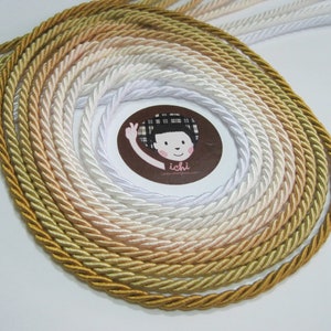 Puede incluir: Una colección de cordones trenzados en varios tonos de dorado, crema y blanco. Los cordones están dispuestos en un patrón circular, con el tono más oscuro en el borde exterior y el tono más claro en el centro.