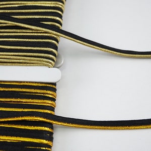 Könnte beinhalten: Schwarzes und goldenes elastisches Band mit einem dünnen goldenen Streifen entlang der Kante. Das Band ist um eine Spule gewickelt.