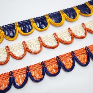 May include: Close-up of three decorative trims with a scalloped edge design. The top trim is navy blue and yellow, the middle trim is off-white and orange, and the bottom trim is orange and navy blue. The trims are on a white background.