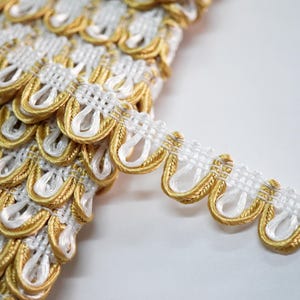 May include: Close-up of decorative trim with a repeating pattern of gold and white scalloped edges. The trim has a woven white base with gold detailing, creating a textured and elegant appearance. Suitable for embellishing garments or craft projects.