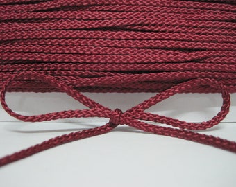 May include: A close-up of a burgundy braided cord tied in a bow. The cord is made of a soft, smooth material and has a subtle sheen.