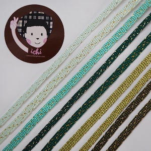 May include: Selection of decorative cords in shades of light blue, turquoise, dark green, and gold. Each cord has a woven pattern with metallic detailing. A circular sticker with a cartoon character is in the top left corner.