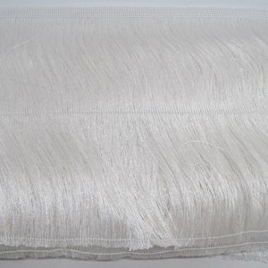 May include: Close-up of white fringe trim. The trim has a dense, textured fringe hanging from a mesh backing. The fringe is made of long, flowing strands, creating a soft, fluffy appearance. Suitable for various craft and sewing projects.