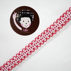 Puede incluir: Cinta decorativa roja y blanca con un diseño de borde festoneado. La cinta se coloca en diagonal a través de la imagen. Una pegatina circular con una ilustración de dibujos animados y el texto "ichi" está en la esquina superior izquierda.