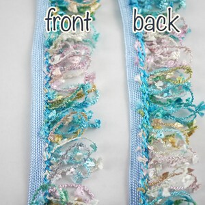 May include: A close-up of light blue trim with a multi-colored fringe. The trim has a woven texture and the fringe is made of various shades of blue, green, pink, and white. The words "front" and "back" are written on the trim.