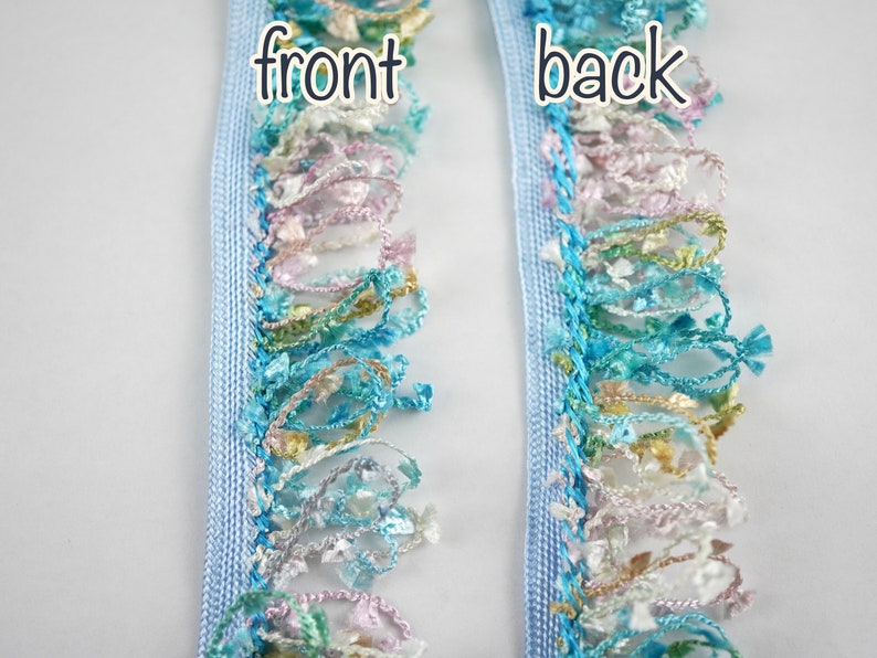 May include: A close-up of light blue trim with a multi-colored fringe. The trim has a woven texture and the fringe is made of various shades of blue, green, pink, and white. The words "front" and "back" are written on the trim.
