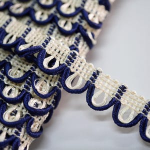 May include: Close-up of decorative trim with a scalloped edge. The trim features a cream-coloured open weave with navy blue edging. The navy blue trim creates a scalloped design. Suitable for sewing and craft projects.
