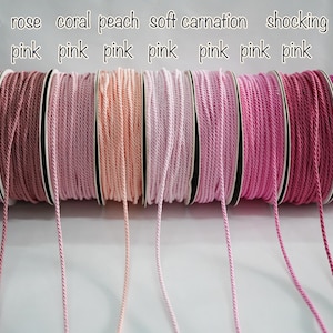 Puede incluir: Se muestran carretes de cordón rosa en varios tonos, desde malva hasta rosa intenso. La imagen muestra una gama de colores, incluyendo rosa, coral, melocotón y rosa suave. El texto en la parte superior identifica cada color.