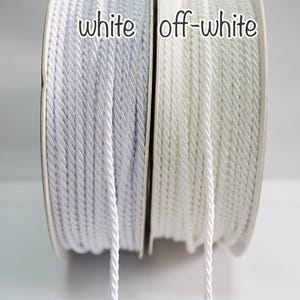 Könnte beinhalten: Zwei Spulen mit gedrehtem Kordel, eine weiße und eine cremefarbene, werden gezeigt. Die Wörter "white" und "off-white" sind über jeder Spule aufgedruckt. Die Kordel ist eng auf die Spulen gewickelt und zeigt ihre Textur und Farbvariationen.