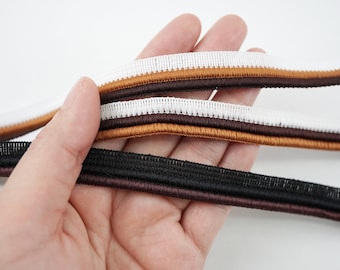 Könnte beinhalten: Nahaufnahme von mehreren elastischen Bändern in verschiedenen Farben. Die Bänder umfassen Weiß, Braun und Schwarz, jedes mit einem kontrastierenden Streifen. Die Bänder werden in einer Hand vor weißem Hintergrund gehalten. Diese Bänder eignen sich für Näh- und Bastelprojekte.