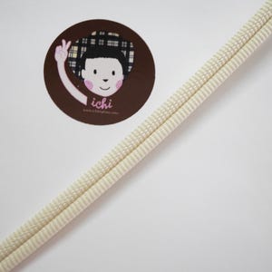 May include: Cream-colored, double-corded trim on a white background. A circular brown sticker with a cartoon illustration of a person with a plaid hat and the text "ichi" is in the upper left corner.