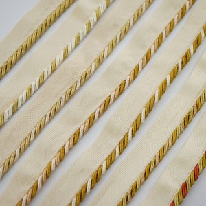 Könnte beinhalten: Fünf verschiedene Borten aus beigefarbenem Stoff mit goldenen, weißen und orangen Kordelverzierungen. Die Borten sind in Reihen angeordnet, wobei die obere Reihe die breiteste und die untere Reihe die schmalste Borte hat.