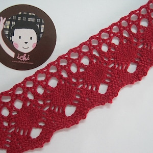 Puede incluir: Un trozo de encaje rojo tejido a ganchillo con un borde festoneado. El encaje presenta un patrón repetitivo de círculos calados y diseños florales. Una pegatina circular con una ilustración de dibujos animados y el texto "ichi" está en la esquina superior izquierda.