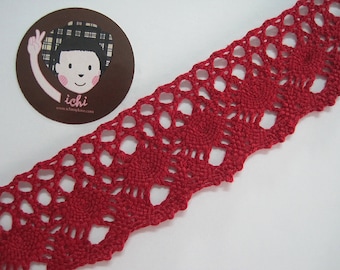 May include: A length of red crocheted lace with a scalloped edge. The lace features a repeating pattern of openwork circles and floral-like designs. A circular sticker with a cartoon illustration and the text "ichi" is in the upper left corner.