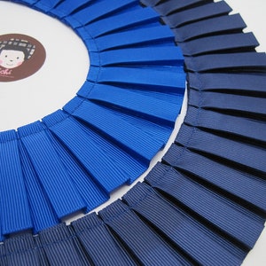 Puede incluir: Dos rollos de cinta plisada, uno azul real y otro azul marino. La cinta está doblada en círculo y los pliegues miran hacia afuera.