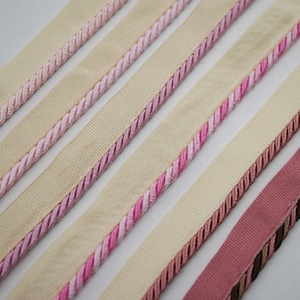 Könnte beinhalten: Fünf verschiedene Borten aus rosa und beigefarbenem Stoff mit einer seilartigen Textur. Die Borten sind in Reihen angeordnet, wobei die rosa Borte oben und die beige Borte unten liegt.