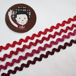 Puede incluir: Cuatro filas de cinta ondulada roja, rosa y burdeos. Una pegatina redonda marrón con una ilustración de dibujos animados de una persona con la palabra "ichi" está en la esquina superior izquierda. La cinta probablemente se utiliza para proyectos de artesanía o costura.