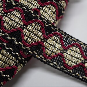 May include: Close-up of decorative trim with a black woven base, gold metallic threads, and red stitching. The trim has a repeating pattern and is suitable for embellishing clothing or craft projects.
