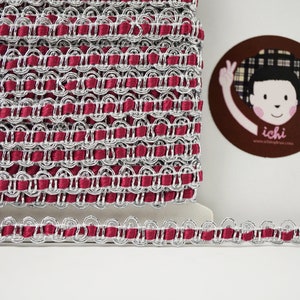 May include: A spool of silver and burgundy trim with a decorative scalloped edge. The trim is made of a woven fabric with a metallic thread.