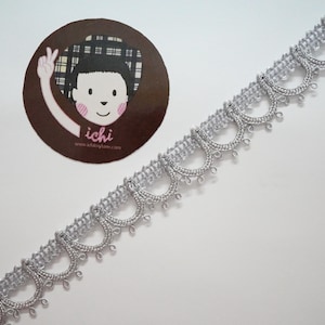 Puede incluir: Adorno plateado con borde festoneado y pequeñas cuentas que cuelgan de la parte inferior. El adorno está sobre un fondo blanco.