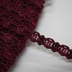 May include: A deep burgundy gimp trim featuring a repeating decorative pattern of looped cord and vertical thread bars. The trim is coiled in a dense stack, with one length extending outward to display the intricate woven texture and dark red colour.