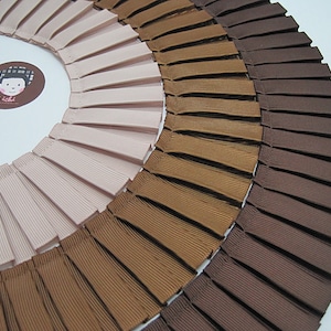 May include: A close-up of three rows of pleated ribbon trim in pink, brown, and dark brown. The ribbon is arranged in a semi-circle.