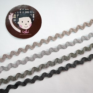 May include: Four strips of wavy ribbon in shades of brown, grey, and black. A circular sticker with a cartoon illustration of a person giving a peace sign is in the upper left corner. The sticker has the word "ichi" on it.