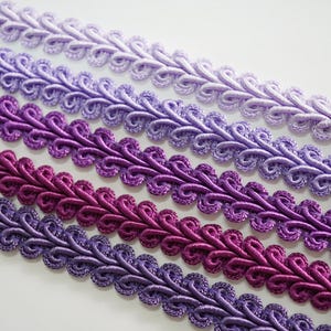 May include: Close-up of several rows of decorative trim in shades of purple. The trim has a looped design, with colors ranging from light lavender to deep magenta. The texture appears to be smooth and the trim is likely made of fabric.