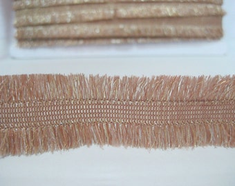 May include: A close-up of a beige and gold fringe trim. The trim has a woven center and a fringe on both sides.