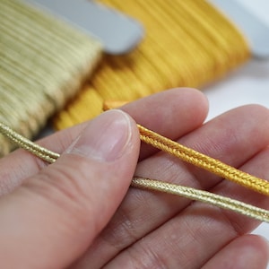 Puede incluir: Dos carretes de cordón metálico, uno en oro pálido y otro en oro amarillo vibrante. Cada carrete contiene una larga longitud de cinta trenzada. Los cordones tienen una textura brillante y reflectante y se muestran como hebras individuales.