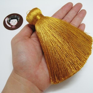 Könnte beinhalten: Ein goldener Quaste mit einem langen, fließenden Fransen. Die Quaste wird in einer Hand gehalten, wodurch Größe und Textur deutlich werden.