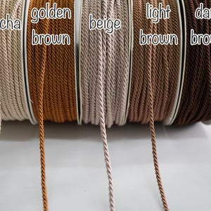 Può includere: Bobine di cordoncino ritorto in varie tonalità di marrone, tra cui moka, marrone dorato, beige, marrone chiaro e marrone scuro. Ogni bobina è etichettata con il nome del colore in testo bianco. I cordoncini sono ben avvolti e sembrano essere fatti di un materiale liscio e resistente.