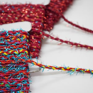 Puede incluir: Primer plano de carretes de cinta decorativa en tonos rojos, azules y amarillos. La cinta tiene un aspecto texturizado y tejido con acentos metálicos. La imagen muestra los colores vibrantes y los detalles intrincados de la cinta, adecuada para manualidades.